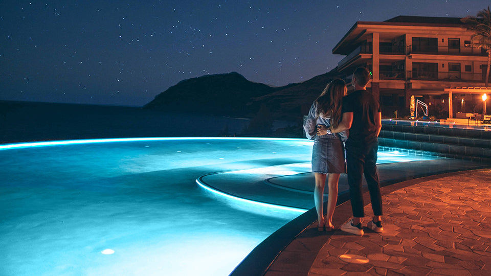 Two people standing by the swimming pool with pool lights on